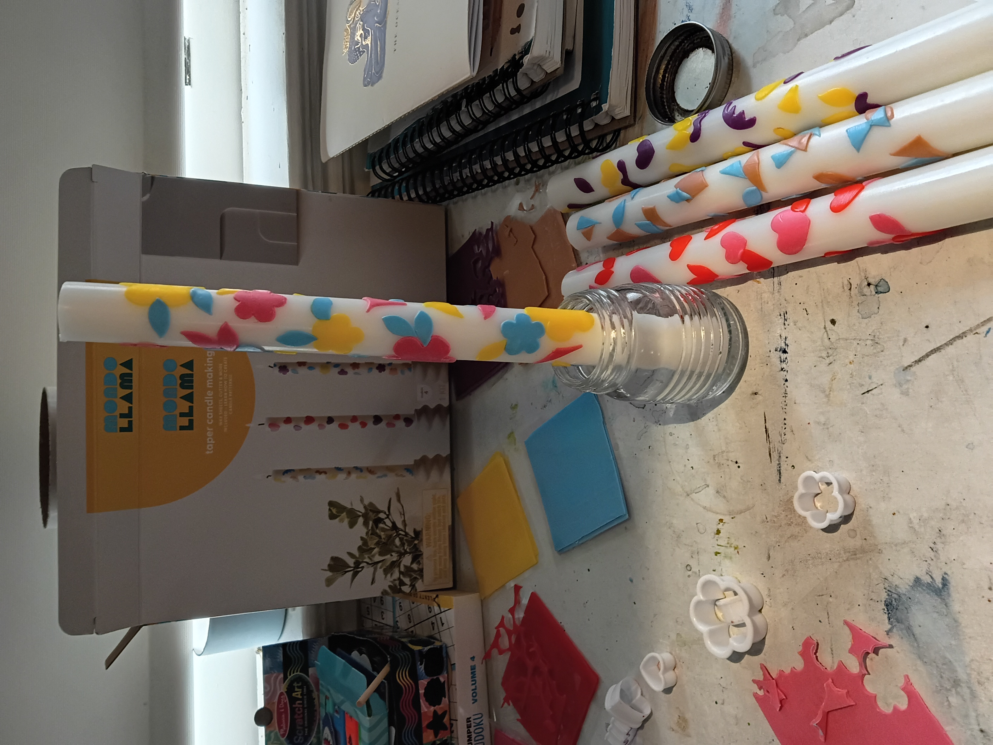 a cluttered desk with a set of four candles with different colored patterns, the front candle is resting in a mini mason jar the rest are laying down